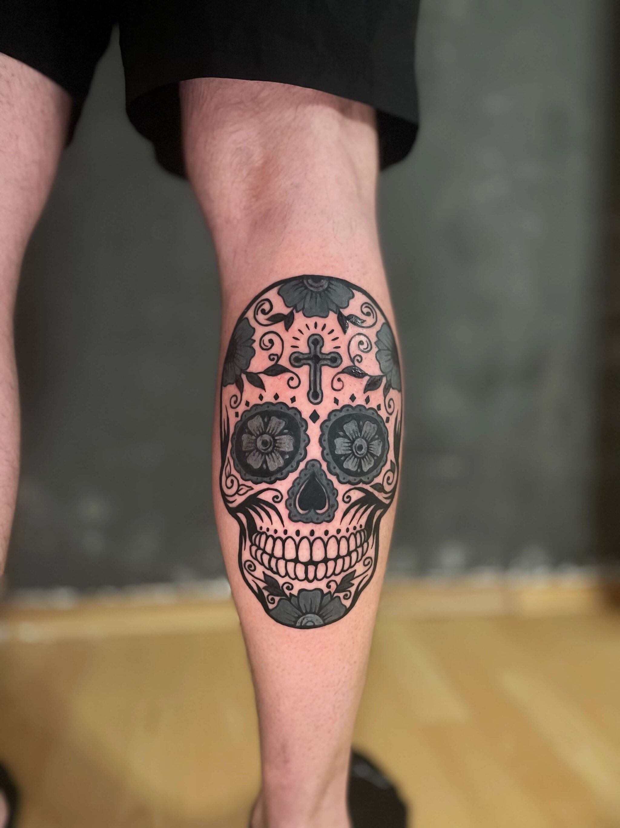 Sugar Skull Tattoo auf der Wade Black and Grey Sugar Skull Tattoo auf der Wade mit floralen Details und Kreuz – gestochen von Roman Schürr Tattoo
